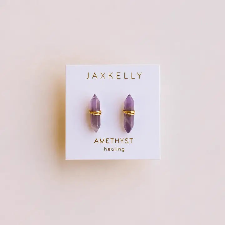 A pair of gold wire-wrapped amethyst stud earrings from JaxKelly, shown on a white card reading "Amethyst Mineral Point - Healing" against a beige background. These gold-plated, tarnish-free earrings are ideal for daily wear.