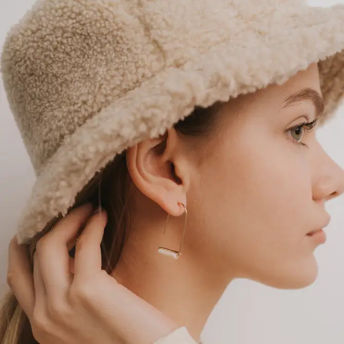 A young woman in a fuzzy beige bucket hat looks to the side, touching her ear. She wears JaxKelly's Mother of Pearl Gemstone Drop - Calming earrings, with her light brown hair tucked behind her ear.