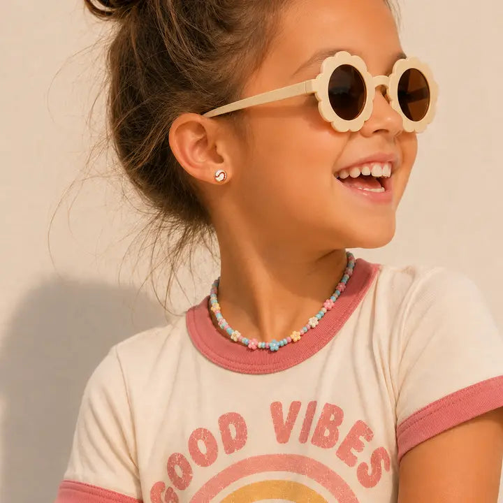 A smiling child wearing flower-shaped sunglasses, a pastel bead necklace, and a shirt with pink trim that says GOOD VIBES shows off JaxKelly’s Softball Enamel Flat Back Kids Earrings—fun, hypoallergenic accessories crafted from surgical-grade stainless steel.