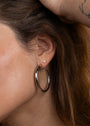 A close-up shows a person wearing the Silver Everyday Hoop - Large in sterling silver, paired with a small stud earring. A hand gently touches their brown, wavy hair above the ear, highlighting this timeless jewelry piece.