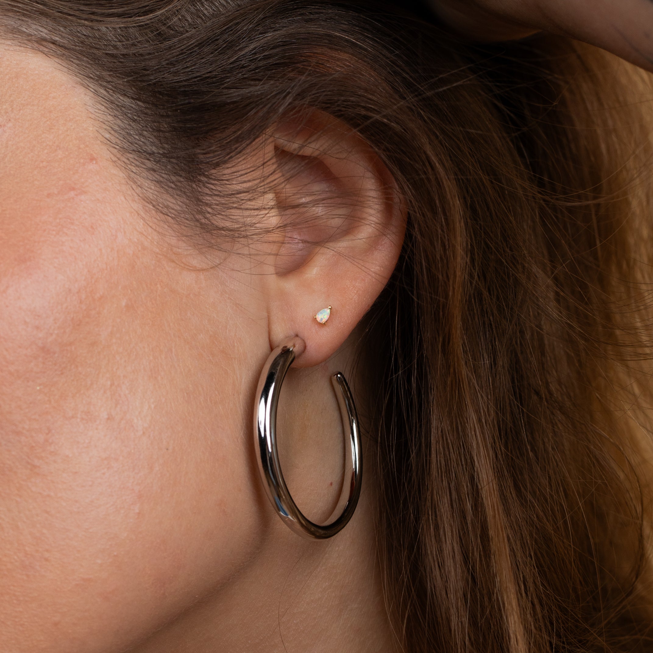 A close-up shows a person wearing the Silver Everyday Hoop - Large in sterling silver, paired with a small stud earring. A hand gently touches their brown, wavy hair above the ear, highlighting this timeless jewelry piece.
