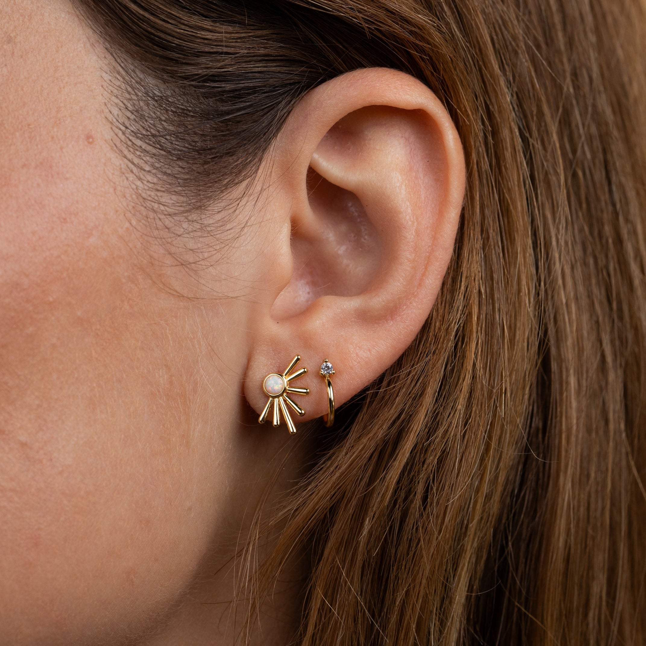 Close-up of a woman's ear with straight, light brown hair wearing two gold earrings: the Sun Ray - White Opal earring featuring a lab-grown White Opal, and a small 18k gold plated hoop.