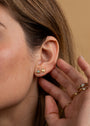 A close-up of a person’s ear wearing the Amazonite Mini Energy Gems - Hope: two gold earrings—a sun and a small amazonite triangle—with sterling silver posts and 18k gold plating. Their hand, with elegant gold rings, gently touches their ear.