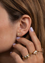 Close-up of a woman's ear wearing the Open Mini Hoop Earring - Pink and a sun stud. Her pastel purple-manicured hand showcases gold rings with white stones and a sparkling pink CZ stone.