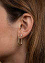 A close-up of a woman's ear shows her brown hair tucked behind, revealing two earrings: a small stud in the upper lobe and the Large Rectangle Hoops—bold, gold, rectangular earrings—in the lower lobe.