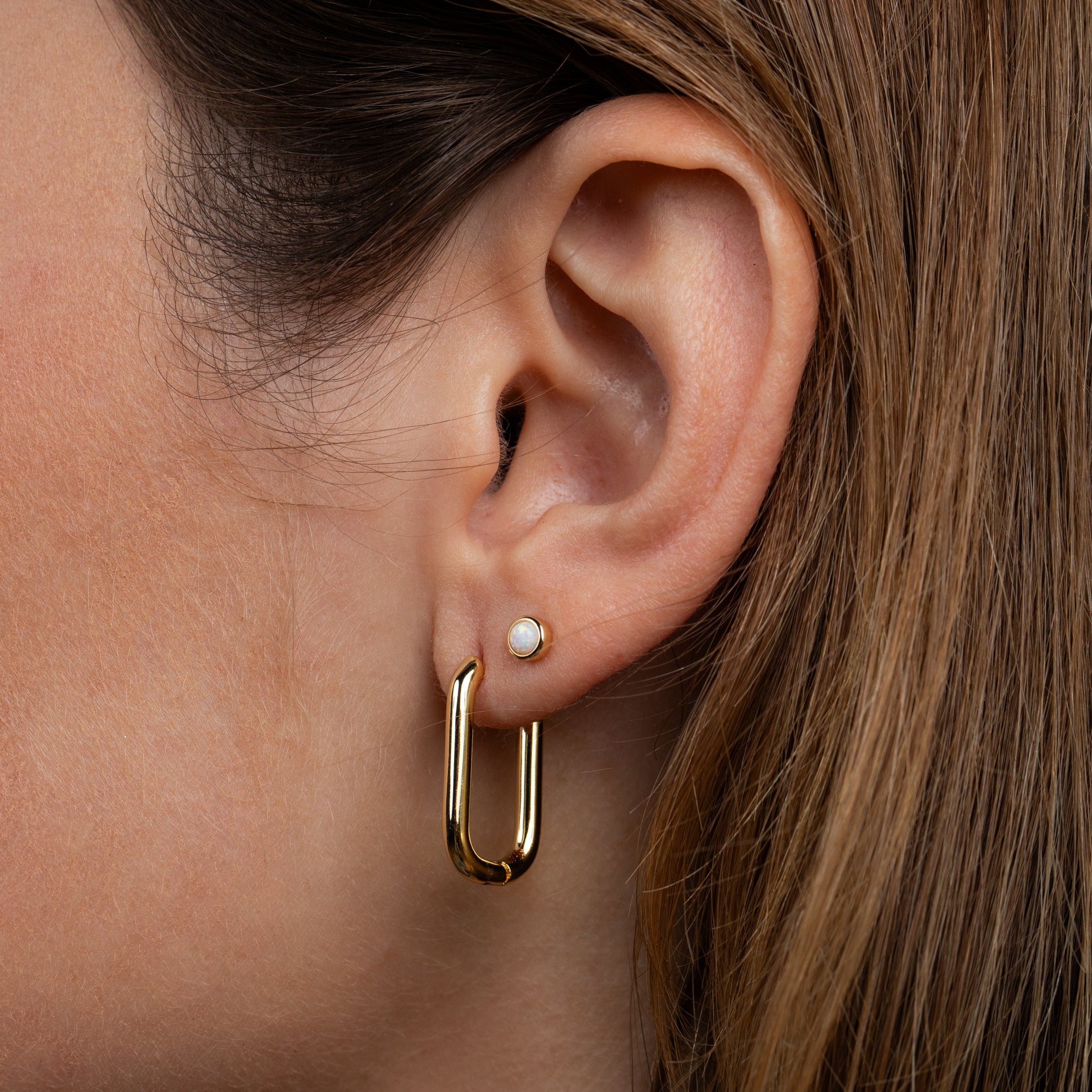 A close-up of a woman's ear shows her brown hair tucked behind, revealing two earrings: a small stud in the upper lobe and the Large Rectangle Hoops—bold, gold, rectangular earrings—in the lower lobe.