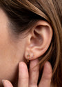 A close-up of a woman's ear with three earrings: the Open Mini Hoop Earring - White (18K gold-plated) in the lobe, a tiny clear stud above it, and a small pink stud beside it. She gently touches her ear with her fingers.