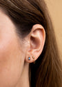 Close-up of a woman’s ear with three earrings: a small gold plated hoop, a round clear sterling silver stud, and the Sapphire Baguette—a rectangular blue stud. Her brown hair is partially visible against a soft beige background.
