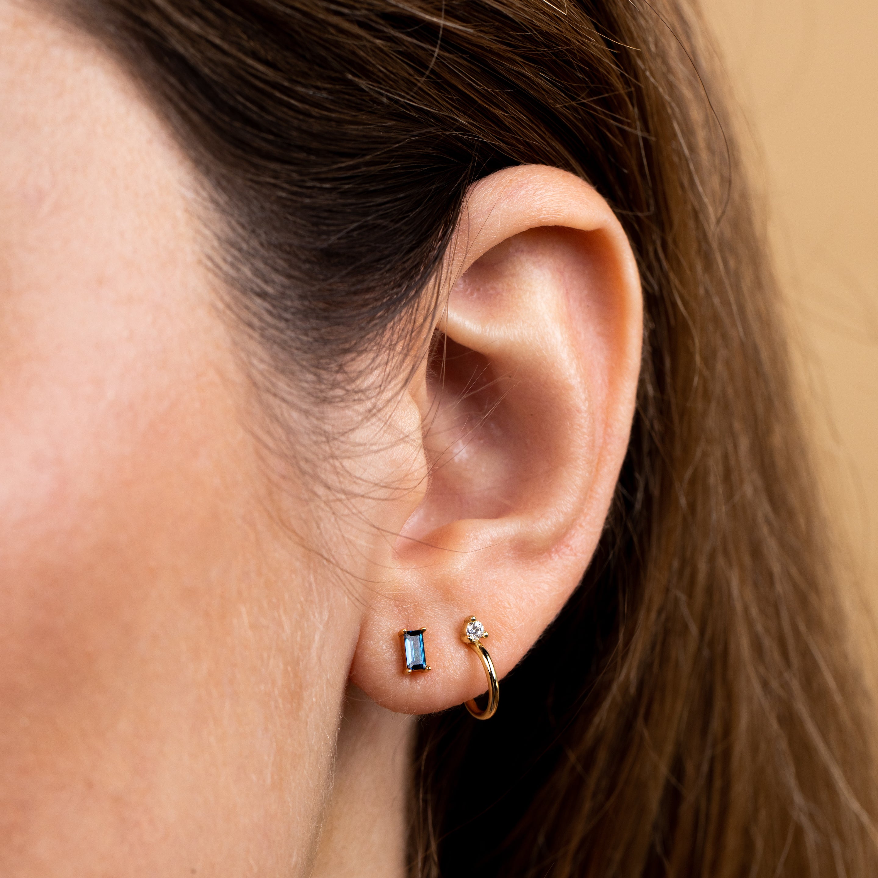 Close-up of a woman’s ear with three earrings: a small gold plated hoop, a round clear sterling silver stud, and the Sapphire Baguette—a rectangular blue stud. Her brown hair is partially visible against a soft beige background.