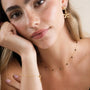 A woman with long blonde hair, calm and relaxed, rests her chin on her hand while wearing JaxKelly’s Double Daisy Drop Earrings, a gold necklace with black beads, and a matching gold-plated bracelet.