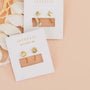 Two pairs of JaxKelly Star Huggie earrings, featuring a gold starburst design with small clear stones, are displayed on white branded cards against a peach background with white textured fabric in the corner.