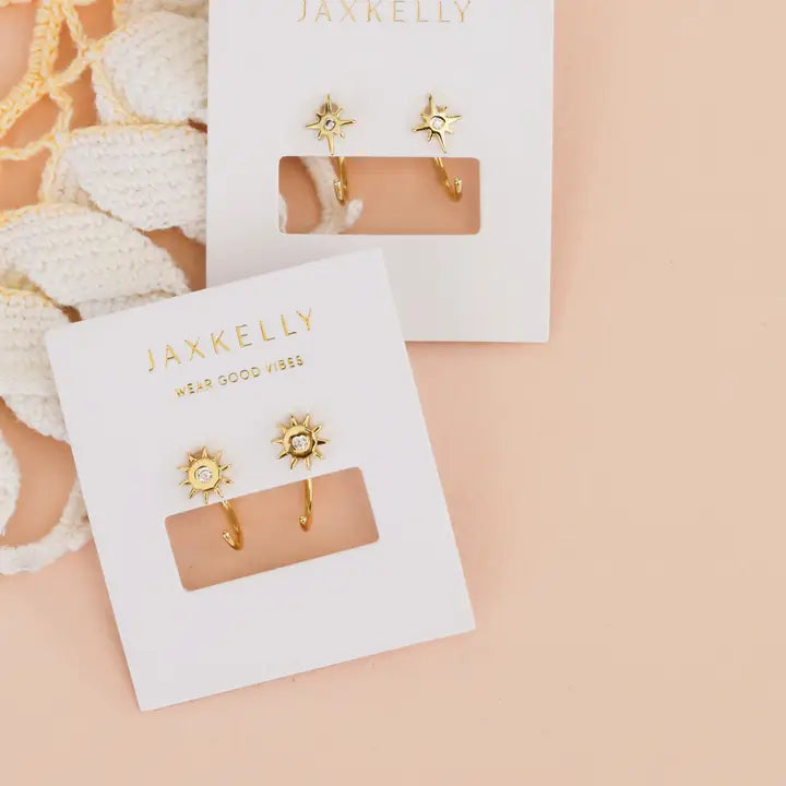 Two pairs of JaxKelly Star Huggie earrings, featuring a gold starburst design with small clear stones, are displayed on white branded cards against a peach background with white textured fabric in the corner.