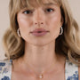 A woman with blonde hair wears JaxKelly Multi Pearl Hoop Earrings and a gold rectangular pendant necklace. She has a neutral expression and is dressed in a floral top against a plain background.