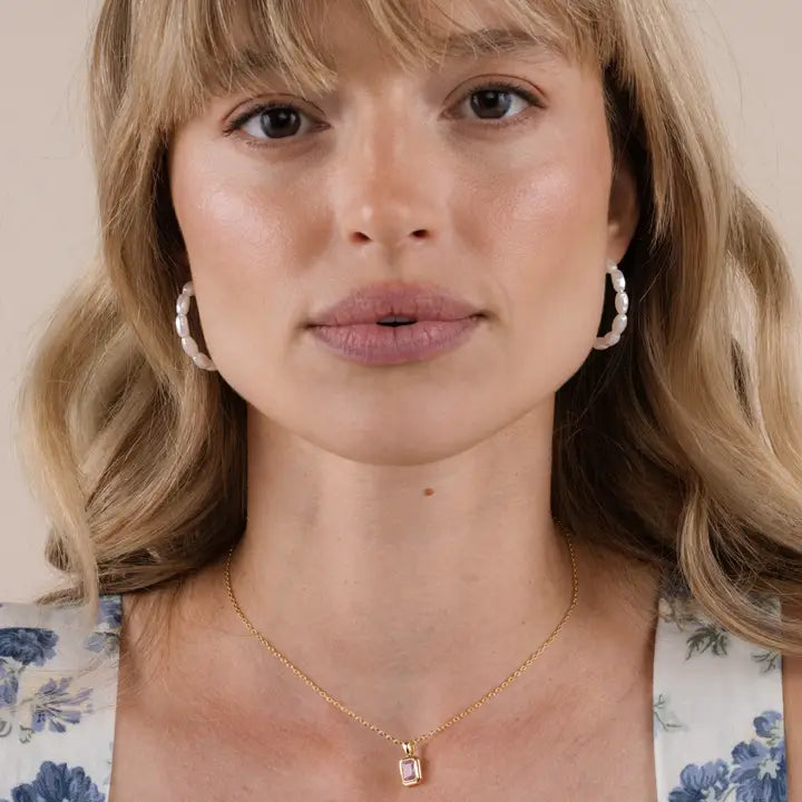 A woman with blonde hair wears JaxKelly Multi Pearl Hoop Earrings and a gold rectangular pendant necklace. She has a neutral expression and is dressed in a floral top against a plain background.