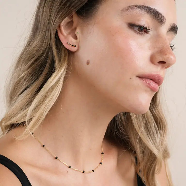 A woman with long blonde hair and clear skin wears a black top, a delicate gold necklace with black beads, and multiple gold-plated earrings—including the striking Black Crawler ear climber from JaxKelly. With prominent eyebrows, she gazes slightly to the side against a neutral background. Her elegant look is completed by her tarnish-free jewelry.