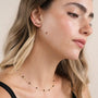 A woman with long blonde hair and clear skin wears a black top, a delicate gold necklace with black beads, and multiple gold-plated earrings—including the striking Black Crawler ear climber from JaxKelly. With prominent eyebrows, she gazes slightly to the side against a neutral background. Her elegant look is completed by her tarnish-free jewelry.