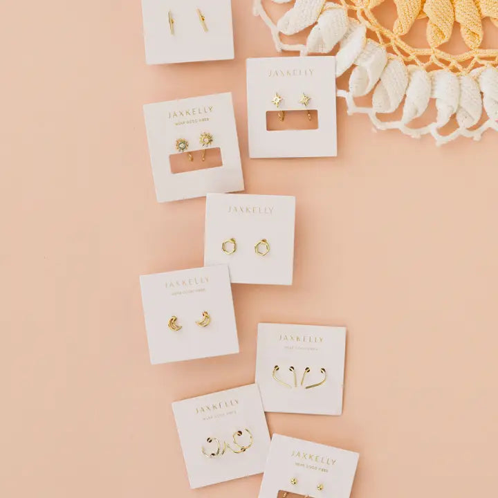 Eight pairs of gold earrings, including the Minimalist Bar by JaxKelly, are displayed on white JaxKelly cards atop a peach background near a crochet doily. These hypoallergenic earrings feature simple, delicate designs.