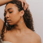 A woman with curly brown hair poses with one hand on her head, wearing JaxKelly's Amazonite Hoop - Hope earrings: large gold hoops featuring a vertical turquoise amazonite pendant, set against a plain light background.
