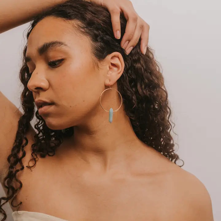 A woman with curly brown hair poses with one hand on her head, wearing JaxKelly's Amazonite Hoop - Hope earrings: large gold hoops featuring a vertical turquoise amazonite pendant, set against a plain light background.
