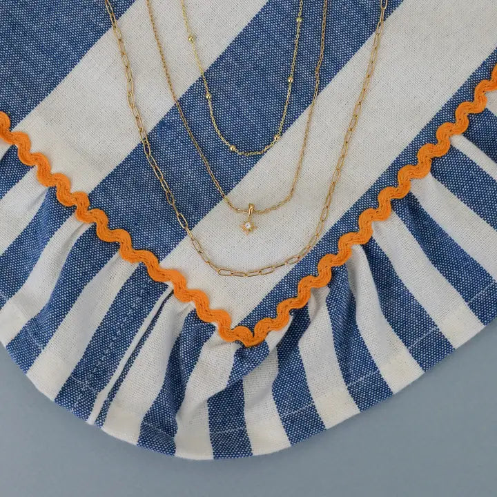The JaxKelly Tiny Star Necklace, plated in 18k gold, is shown on blue and white striped fabric with orange trim. The delicate design features a sparkling Cubic Zirconia pendant and adjustable length for a layered look.