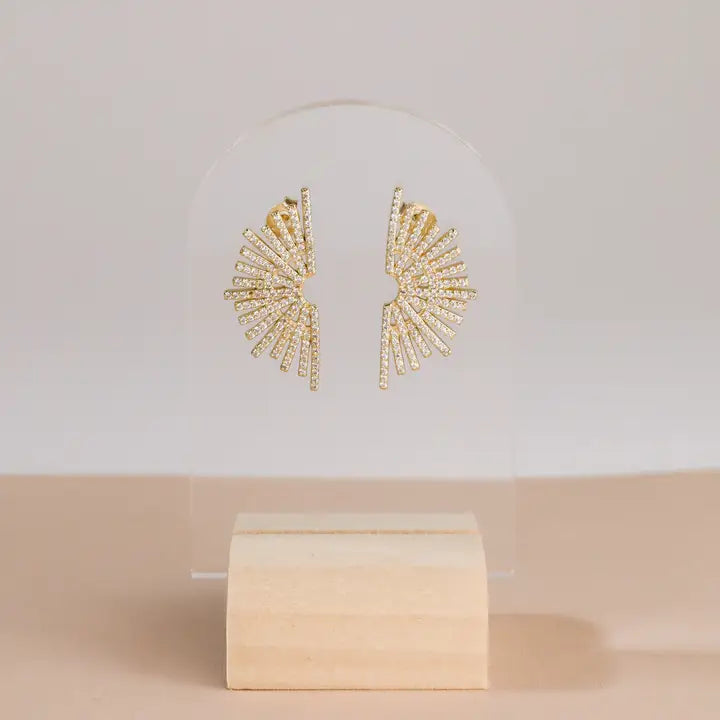 JaxKelly Sunburst Studs, crafted in gold vermeil and adorned with pavé stones, are showcased on a clear acrylic stand with a light wooden base against a neutral background.