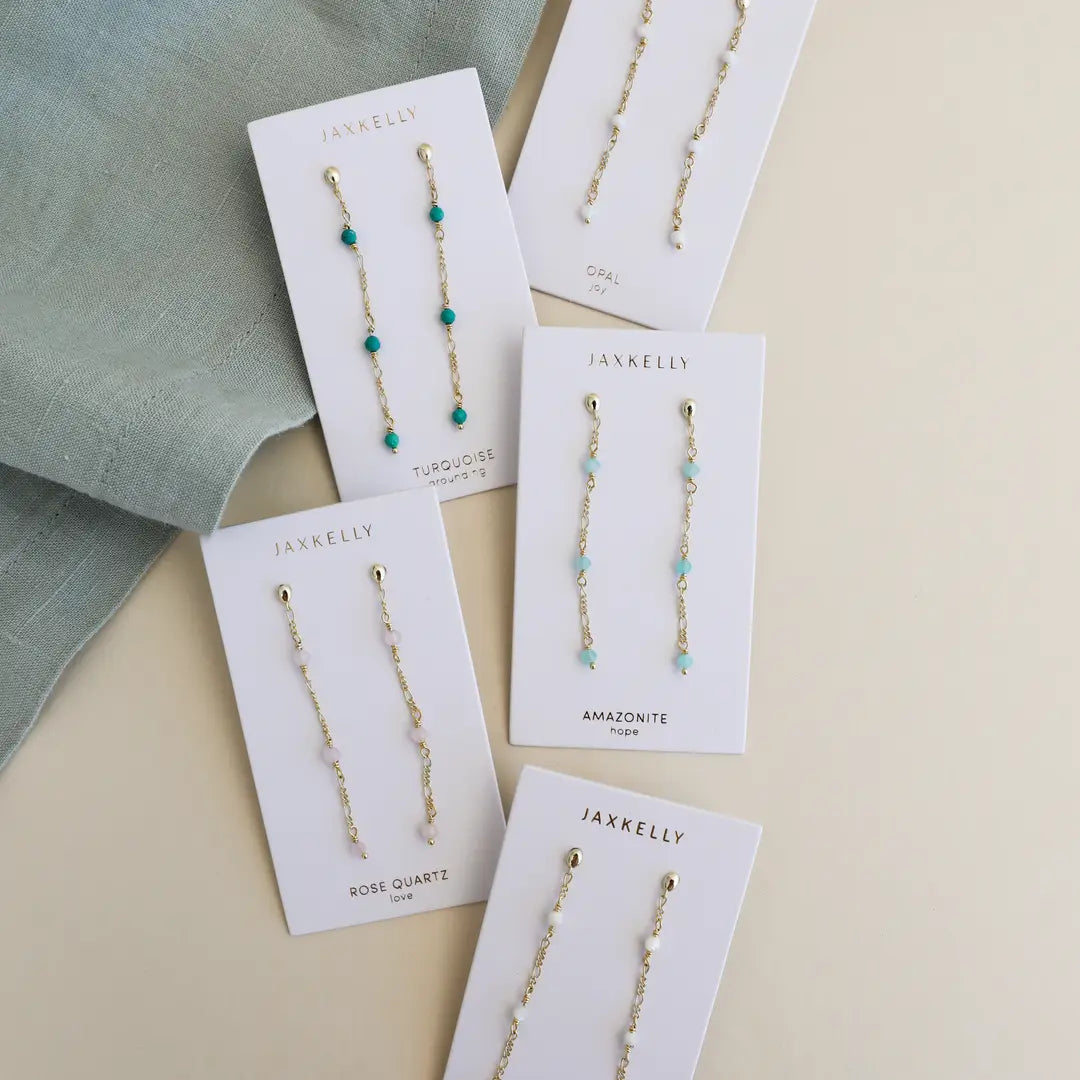 Five pairs of JaxKelly Gemstone Chain Drop - Amazonite earrings with 18k gold plating are displayed on branded cards, featuring Amazonite, turquoise, rose quartz, and opal next to a folded green cloth.