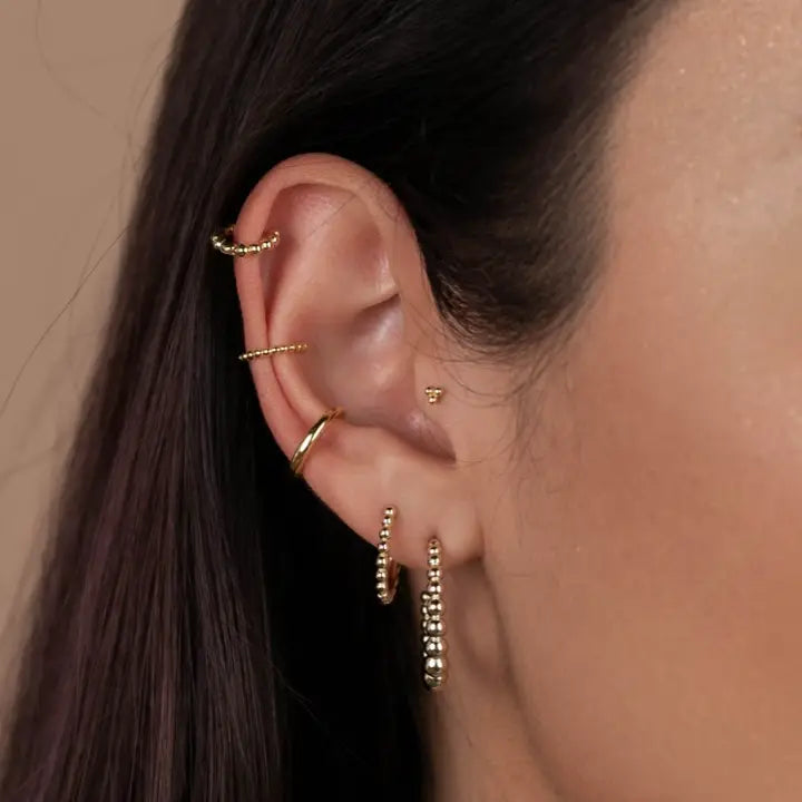 Close-up of a person's ear wearing multiple JaxKelly Small Ball Hoop gold earrings and cuffs, including delicate hypoallergenic styles, with straight dark brown hair tucked behind the ear against a neutral beige background.