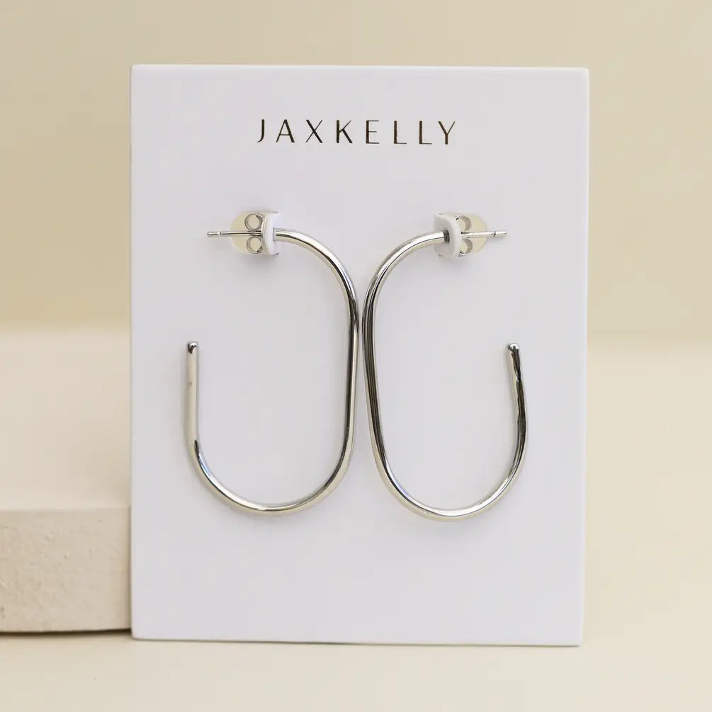 A pair of Silver Large Open Hoop earrings by JaxKelly is shown on a white card with the brand’s name at the top, set against a smooth beige background.