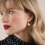 A woman with blonde hair and red lipstick wears a black and white polka-dot top and JaxKelly's Seashell Pearl Drop Earrings, featuring 18K gold plating and a freshwater pearl, as she looks left against a neutral background.