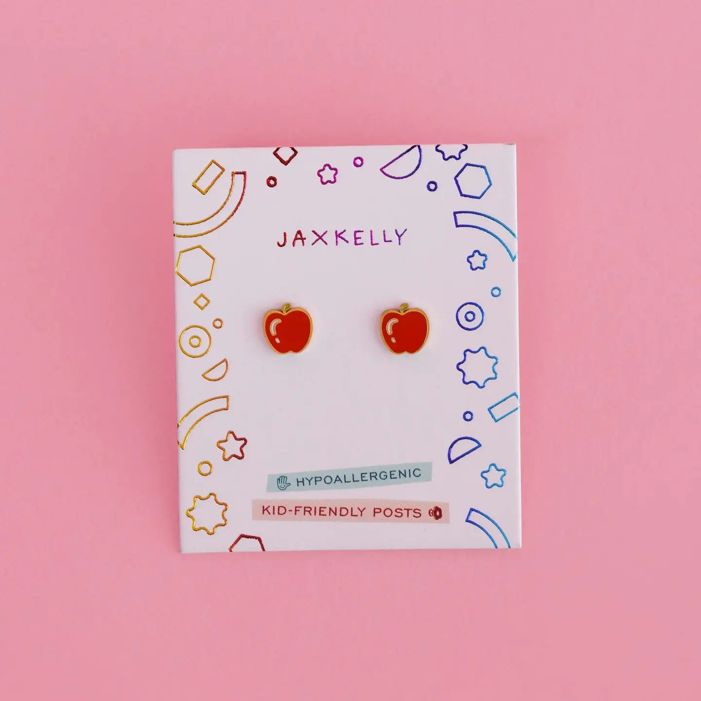 Apple Enamel Flat Back Kids Earrings: small red apple-shaped earrings for kids on a white JaxKelly card with colorful shapes and "hypoallergenic" and "kid-friendly posts" text, made from surgical-grade stainless steel, shown on a pink background.