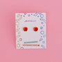 Apple Enamel Flat Back Kids Earrings: small red apple-shaped earrings for kids on a white JaxKelly card with colorful shapes and "hypoallergenic" and "kid-friendly posts" text, made from surgical-grade stainless steel, shown on a pink background.