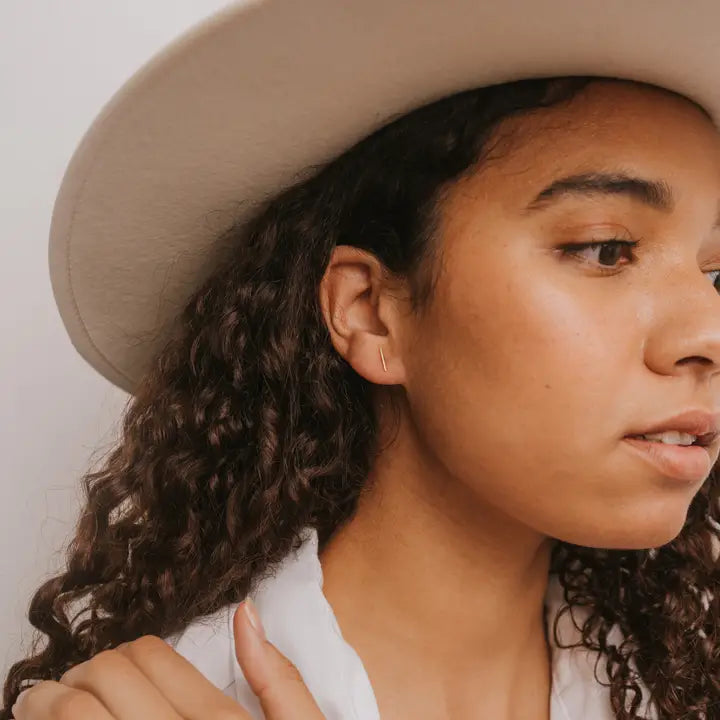 A woman with curly hair in a wide-brimmed hat and white shirt looks to the side, her hand near her collar, subtly showcasing JaxKelly's Minimalist Bar earrings—hypoallergenic and tarnish-free—for an effortless, calm style.