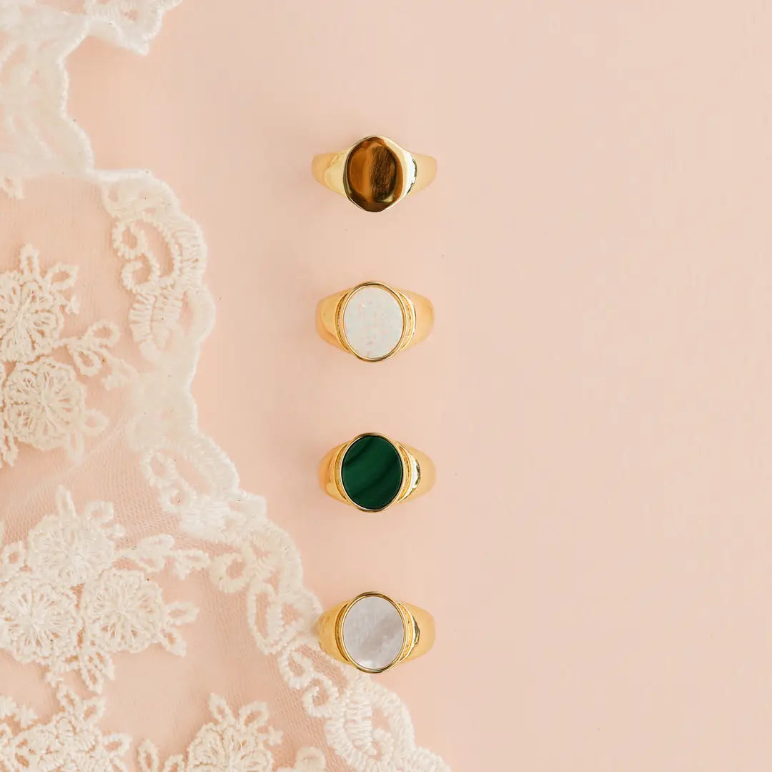 The JaxKelly Mother of Pearl Signet Ring is shown alongside three 18k gold plated rings with brown, white, and green stones, all arranged vertically on a blush pink surface next to delicate white lace fabric.