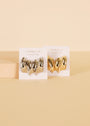 JaxKelly’s Silver Bold Wave chunky hoop earrings with sterling silver posts rest on branded cards—one in silver, one in gold—displayed on a beige surface with a light wooden block behind them.