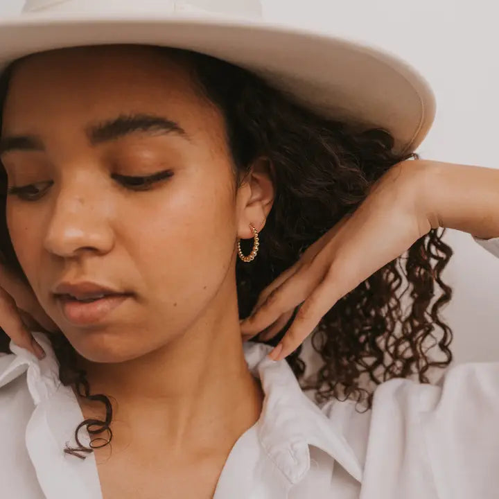 A woman with curly hair wears JaxKelly Large Ball Hoop earrings, a wide-brimmed hat, and a white shirt. She gently touches her neck while looking down against a neutral background.