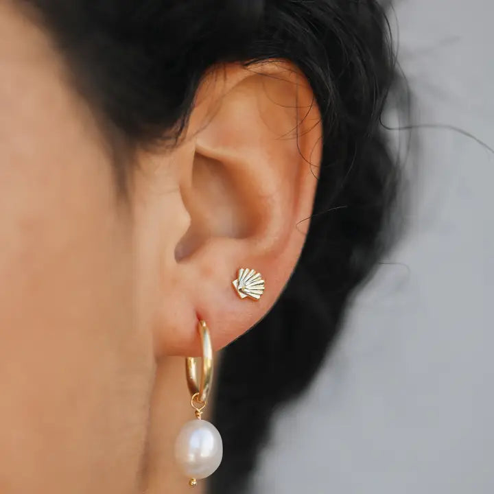 Close-up of an ear wearing the JaxKelly Seashell Stud Earring—tarnish-free gold—with a gold hoop and dangling white pearl. The background is plain and out of focus.