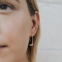 Close-up of a person with light skin and blonde hair wearing JaxKelly's Amazonite Gemstone Drop - Hope: a modern, minimalist 18k gold plated earring featuring a light blue amazonite drop and tubular bead, set against a pale textured background.