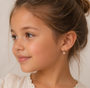 A young girl with light brown hair in a loose bun, wearing a cream lace top and JaxKelly Bow Enamel Charm Hoop earrings, smiles softly to the left against a neutral background.