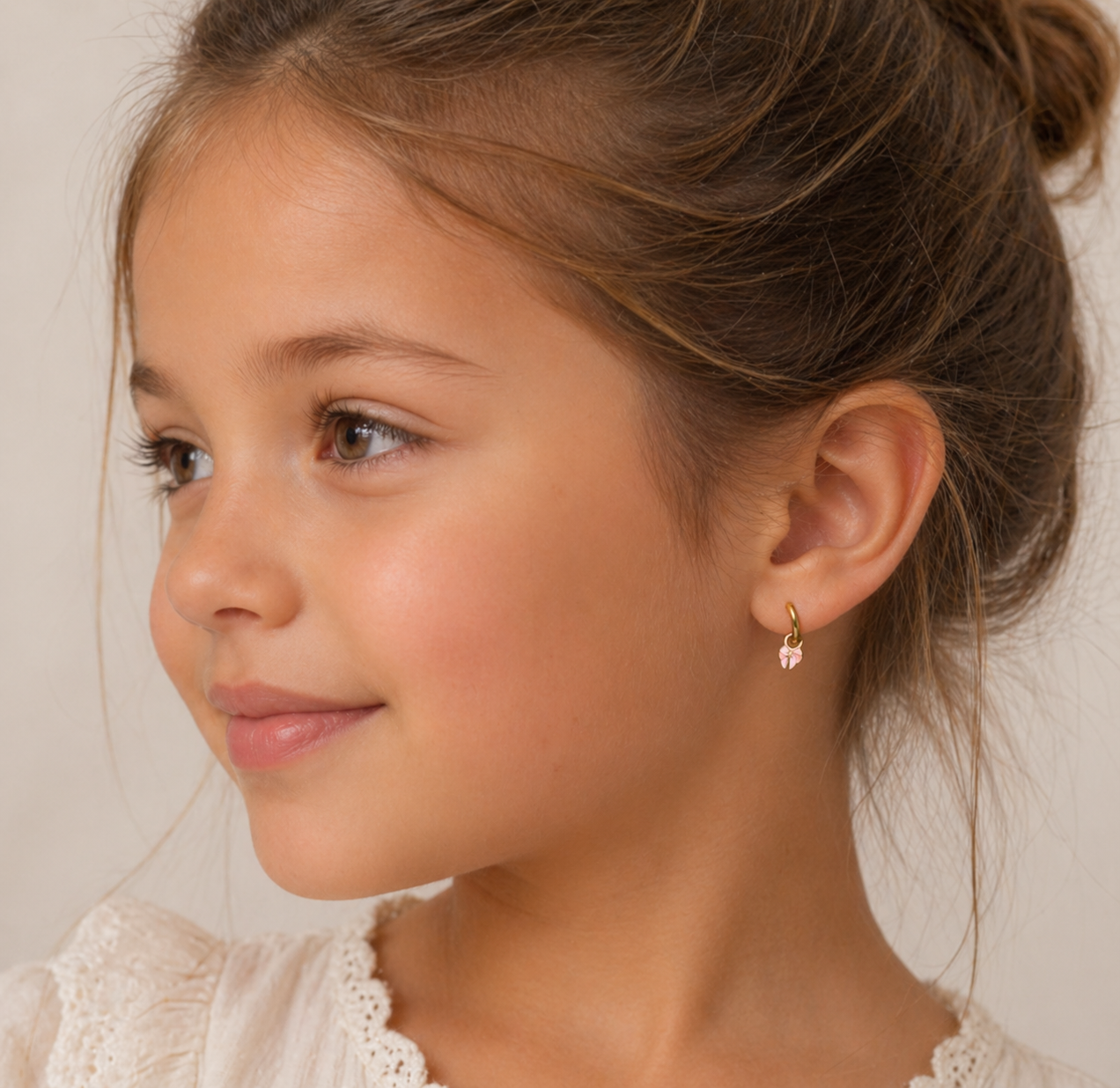A young girl with light brown hair in a loose bun, wearing a cream lace top and JaxKelly Bow Enamel Charm Hoop earrings, smiles softly to the left against a neutral background.