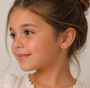 A young girl with light brown hair in a bun, wearing a white lace-trimmed top, smiles softly and gazes to the side. She wears the JaxKelly Watermelon Enamel Charm Hoop, a kids’ earring crafted from surgical grade stainless steel.