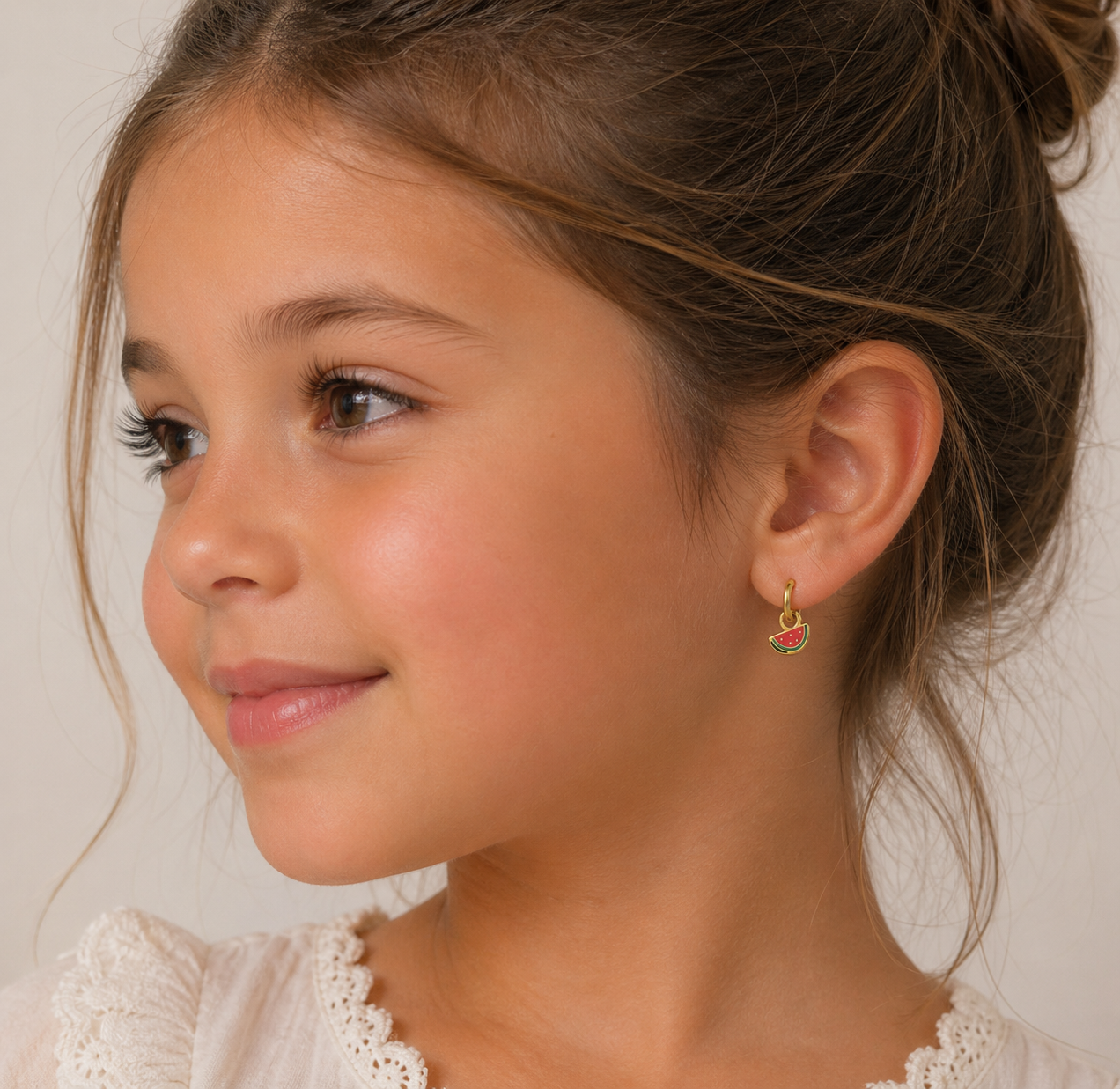 A young girl with light brown hair in a bun, wearing a white lace-trimmed top, smiles softly and gazes to the side. She wears the JaxKelly Watermelon Enamel Charm Hoop, a kids’ earring crafted from surgical grade stainless steel.