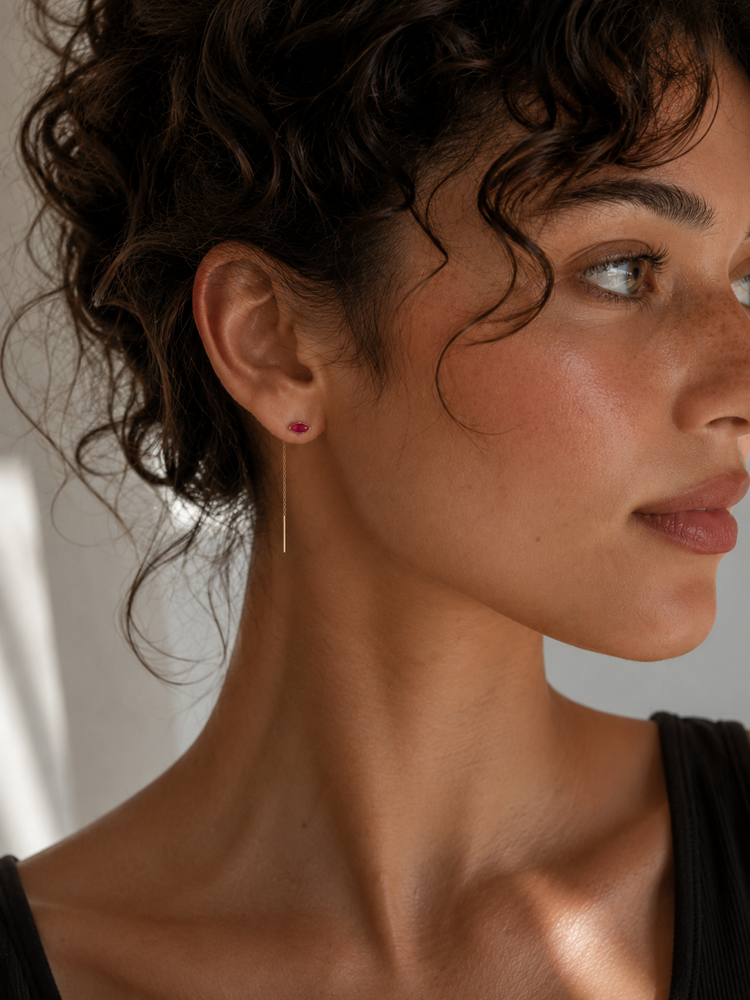 A woman with curly dark hair looks to the right, wearing JaxKelly’s Studded Threader - Pink CZ earring. Her skin glows naturally as soft sunlight highlights her face and neck above a simple black top.