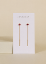 The JaxKelly Studded Threader - Pink CZ earrings feature delicate gold chain threads with pink cubic zirconia studs and 18K gold plating, displayed on a white JaxKelly card against a beige background.