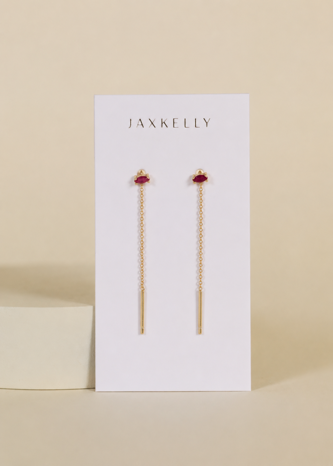 The JaxKelly Studded Threader - Pink CZ earrings feature delicate gold chain threads with pink cubic zirconia studs and 18K gold plating, displayed on a white JaxKelly card against a beige background.