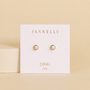 A pair of gold plated, hypoallergenic JaxKelly Simple Stud - White Opal earrings with white opal settings are displayed on a branded card labeled "OPAL joy" against a beige background.