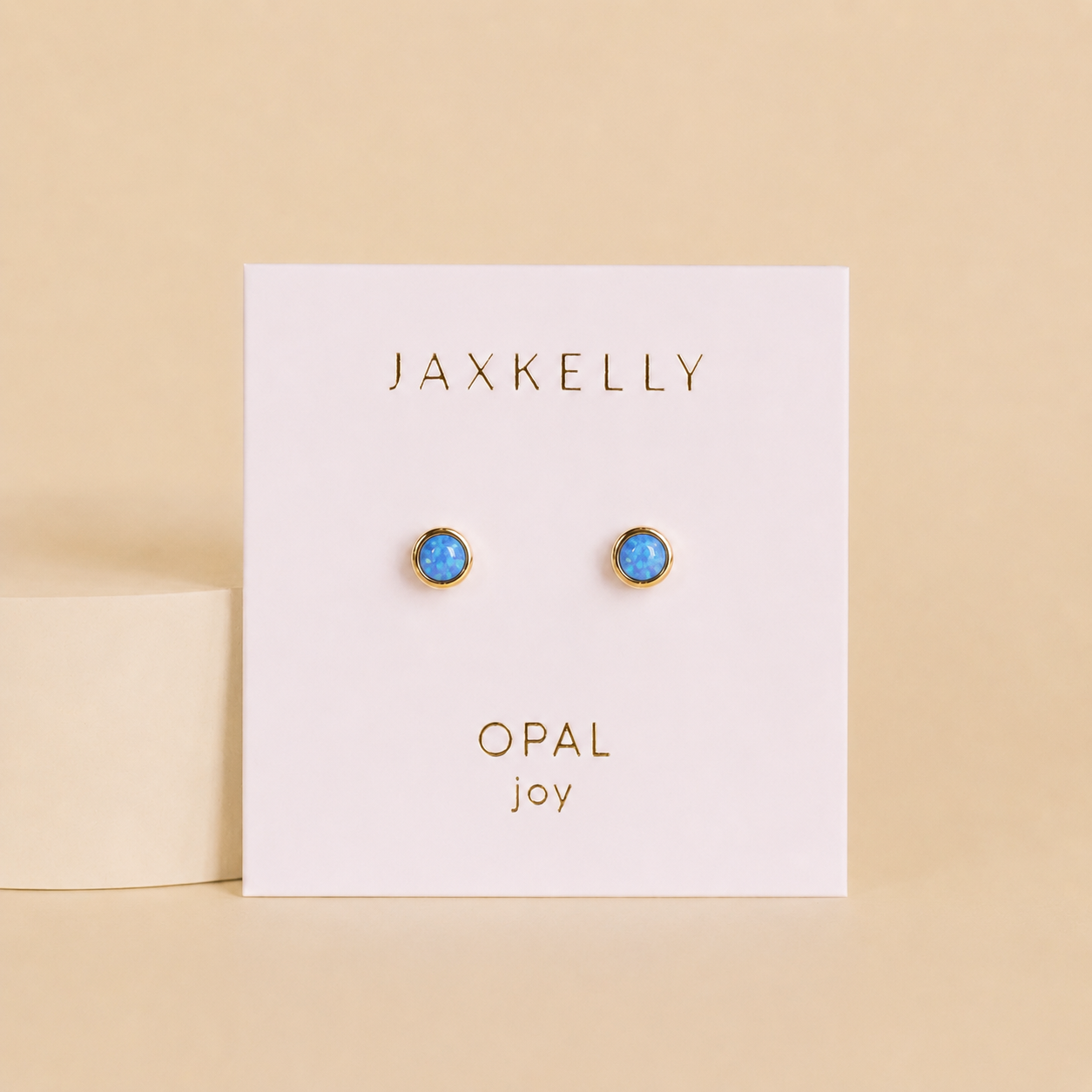 A pair of JaxKelly Simple Stud Earrings in fire opal, gold plated and lab-grown, shown on a white card labeled "OPAL" and "joy" against a beige background.