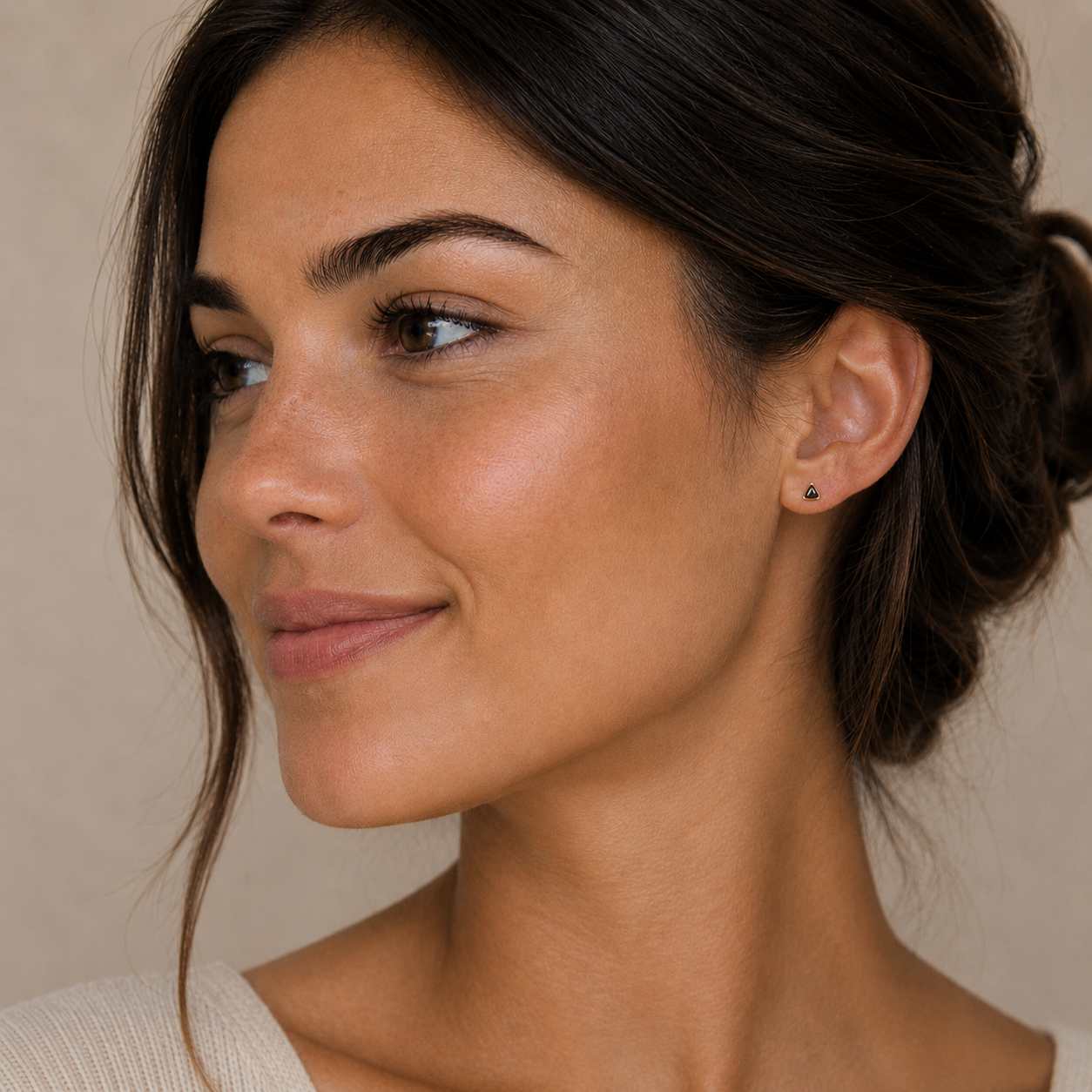 A woman with dark hair in a loose bun smiles softly to the left. She wears JaxKelly Black Tourmaline Mini Energy Gems - Strength studs with sterling silver posts, paired with a beige top against a neutral background.