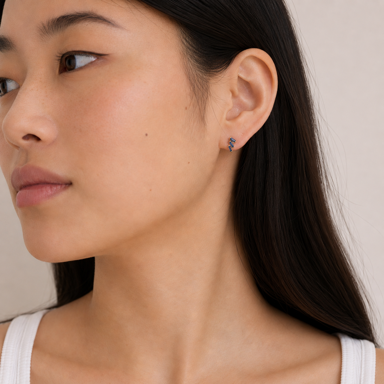 A woman with long dark hair looks to the side, wearing a white tank top and JaxKelly's Offset Trio - Sapphire multicolored hoop earrings with sterling silver posts. The background is neutral beige.