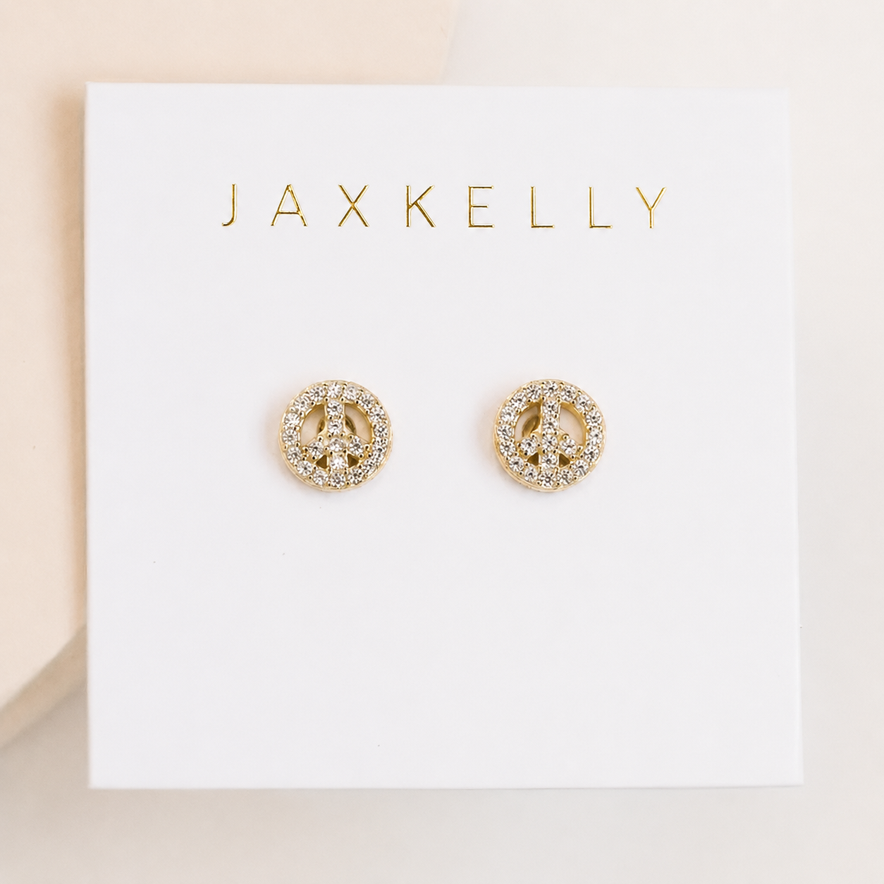 The Pavé Peace Sign earrings by JaxKelly feature sparkling CZ stones and 18k gold plating, displayed on a white card with the JaxKelly logo in gold lettering.