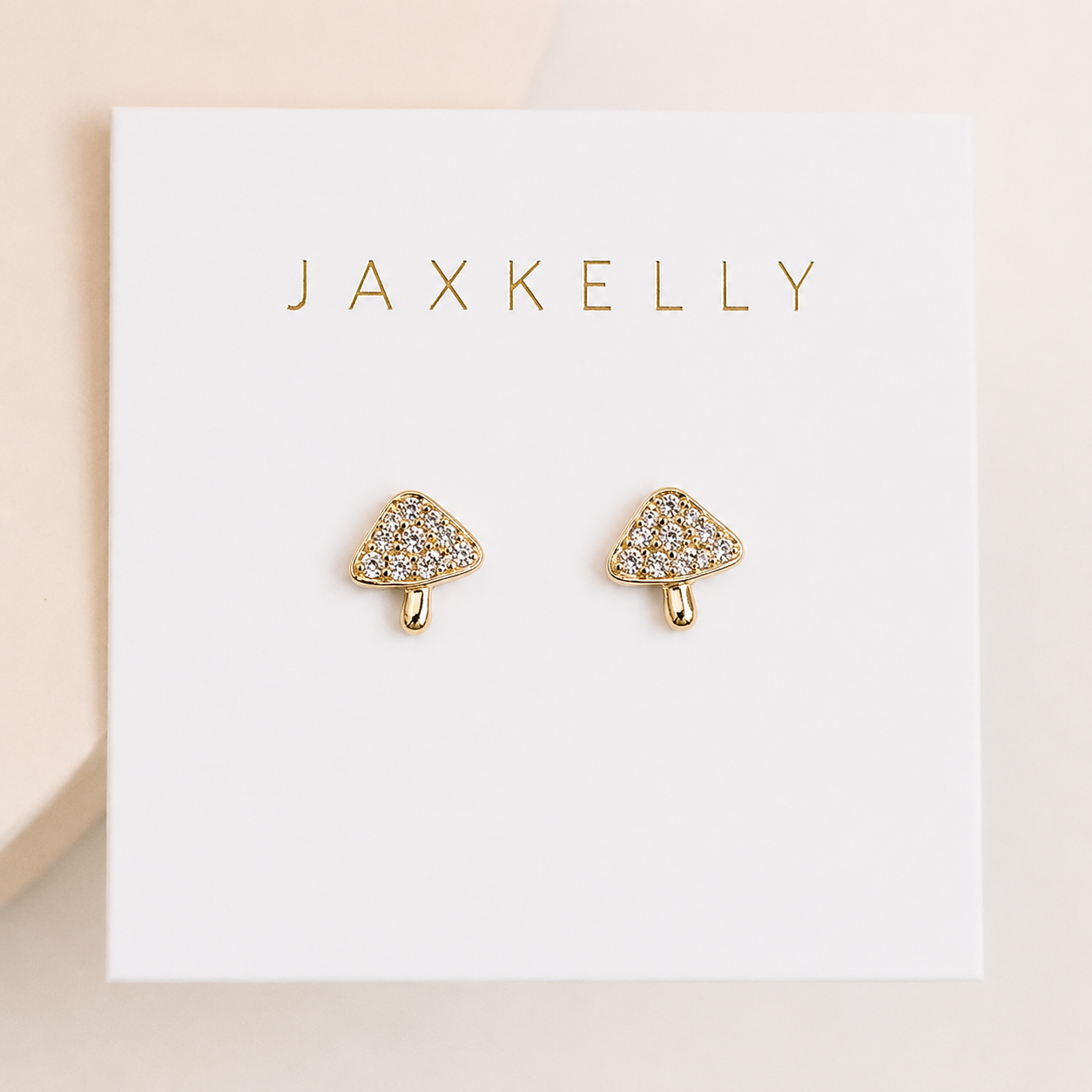 The Pavé Mushroom earrings by JaxKelly feature gold mushroom charms with small clear gemstones and sterling silver posts, displayed on a white card with "JaxKelly" in gold lettering.